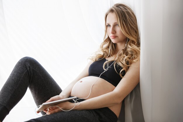 femme enceinte dans des vêtements sportifs sur le rebord de la fenêtre le matin, se détendre, porter des écouteurs, écouter de la musique classique sur tablette numérique.