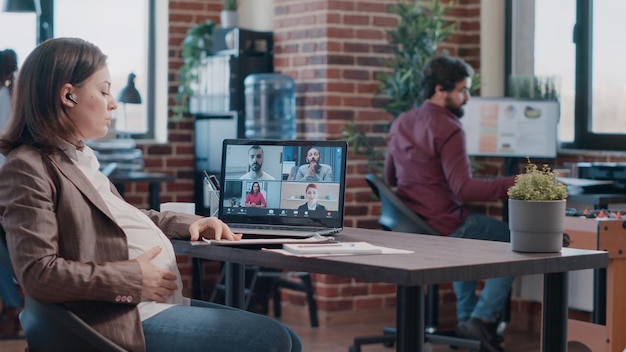 Femme enceinte parlant à des collègues lors d'un appel vidéo, utilisant un ordinateur portable au bureau. L'employé attend un enfant et assiste à une réunion d'affaires avec des collègues en vidéoconférence pour parler du projet