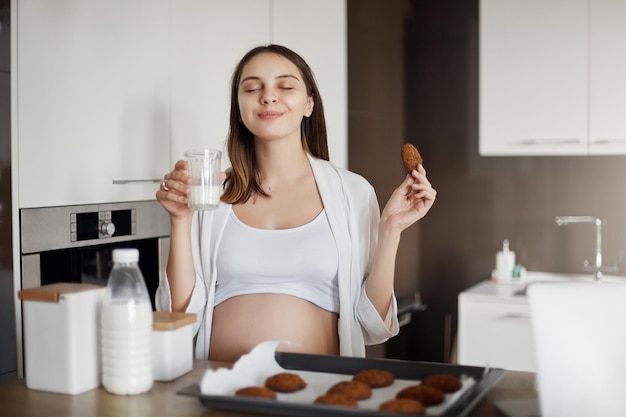 Femme enceinte se sentant ravie et joyeuse appréciant le bon goût de biscuits frais et buvant du lait chaud fermant les yeux et souriant tenant un verre et un biscuit assis près de la table de la cuisine