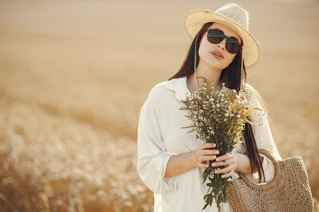 Femme enceinte tenant un bouquet de fleurs sauvages dans un sac de paille, marchant dans un champ de blé. Femme brune marchant dans le champ d'été portant des vêtements blancs