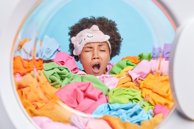 Photo gratuite une femme endormie fatiguée bâille après avoir fait la lessive enfouie dans des vêtements sales multicolores chargés dans la laveuse pour le lavage