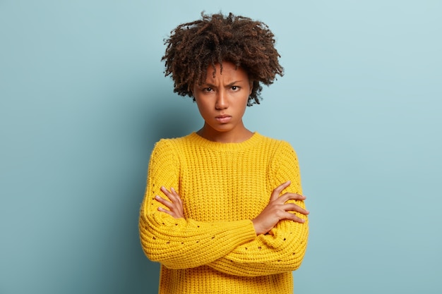 Photo gratuite une femme énervée et énervée croise les mains sur la poitrine, fronce les sourcils et boude, regarde des sourcils, porte un pull jaune tricoté, se tient contre le mur bleu, se sent insultée