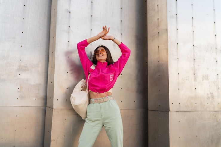 Photo gratuit une femme enjouée s'amusant dans une veste rose néon sur un mur urbain métallique gris