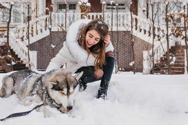 Femme enthousiaste aux cheveux châtain clair regardant son chiot husky et souriant. Portrait en plein air d'une jeune femme heureuse posant avec un chien sur la neige.