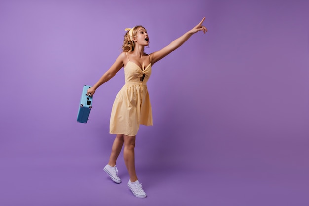 Femme enthousiaste avec valise bleue à la main pointant le doigt vers quelque chose. portrait en pied de drôle de fille curieuse en robe jaune.