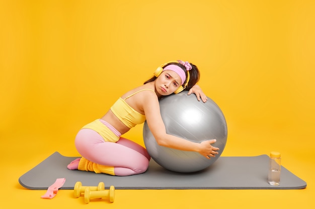 une femme a un entraînement de mise en forme du corps se penche à fitball vêtue de vêtements de sport écoute de la musique via un casque pose sur un tapis de fitness