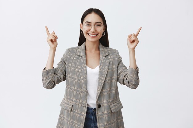 Femme entrepreneur adulte optimiste élégante à lunettes et veste à la mode, levant les mains et pointant vers le haut tout en souriant largement