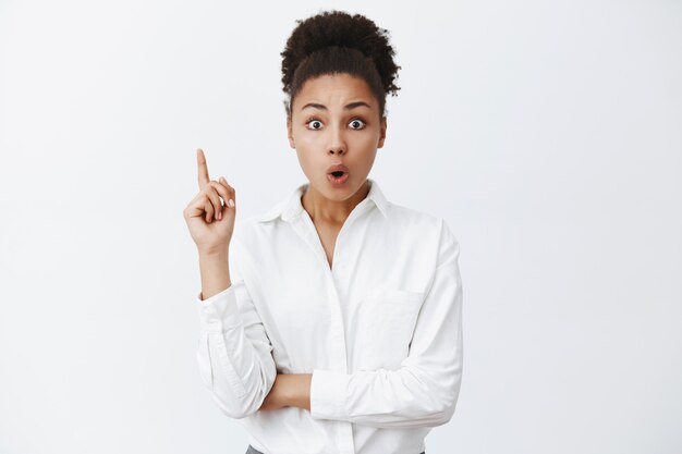 Une femme entrepreneur afro-américaine intelligente et créative en chemise à col blanc, levant l'index dans un geste eureka, pliant les lèvres, ajoutant une excellente suggestion lors d'une réunion d'équipe au bureau, discutant