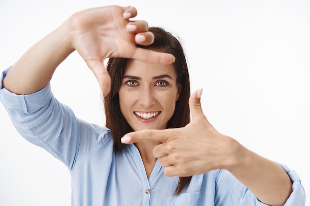 Une femme entrepreneur d'âge moyen inspirée par le gros plan explique sa propre vision, crée une image, montre des cadres de doigt et regarde à travers une scène de prise de vue d'imagerie, se tient debout sur un mur blanc amusé souriant heureux