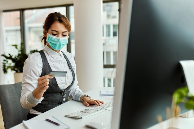 Photo gratuite femme entrepreneur avec masque facial utilisant un ordinateur et une carte de crédit lors de ses achats en ligne au bureau