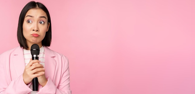 Femme entrepreneur nerveuse prononçant un discours avec microphone tenant un micro et regardant de côté anxieusement posant en costume sur fond rose