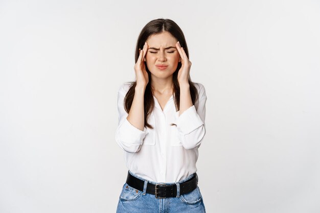 Femme d'entreprise grimaçant, touchant la tête, ressentant des maux de tête, une migraine sévère, debout sur fond blanc