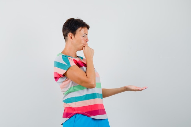 Femme étalant la paume ouverte en t-shirt rayé, pantalon et regardant pensif, vue de face.