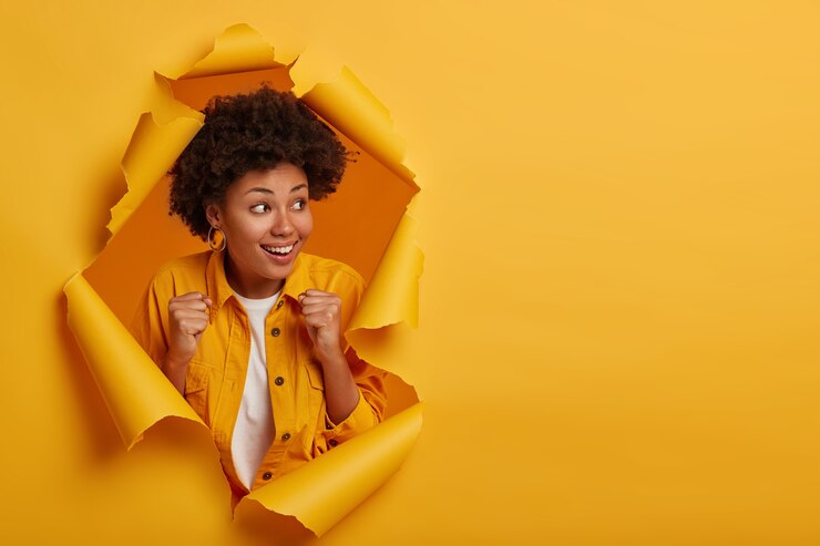 Photo gratuit une femme ethnique joyeuse serre les poings et regarde de côté, se sent très chanceuse et optimiste, célèbre la victoire ou gagne à la loterie, excitée par un résultat positif, se tient dans un trou de papier déchiré.