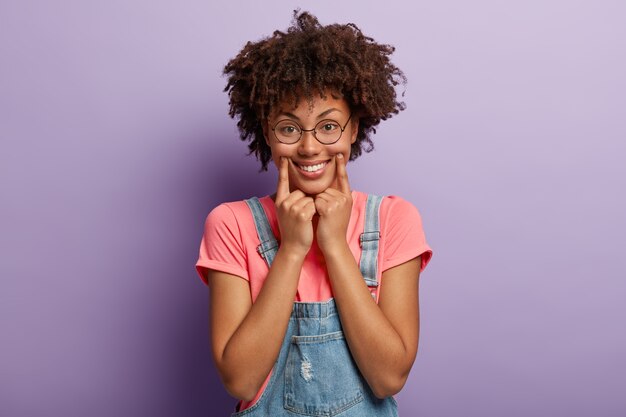 Une femme ethnique joyeuse tient les doigts avant près des coins des lèvres, fait un large sourire, fait semblant d'être de bonne humeur, oublie tous les problèmes, porte des lunettes rondes pour corriger la vue, habillée avec désinvolture