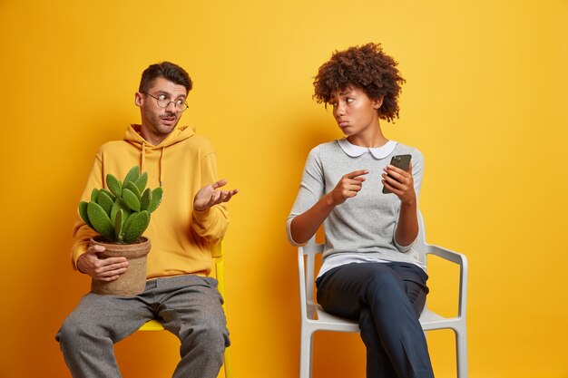 Une femme ethnique mécontente bouleversée indique au smartphone, un homme perplexe hausse les épaules. Un couple interracial tente de résoudre un problème avec un gadget moderne