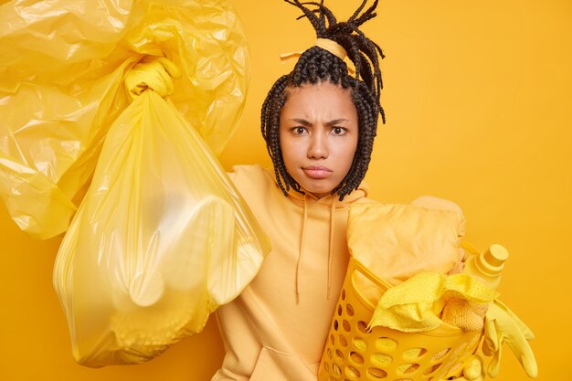 Une femme ethnique mécontente et mécontente avec des dreadlocks porte un sweat-shirt tient un sac poubelle