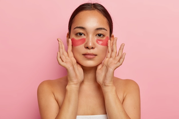 Femme ethnique sérieuse avec des patchs de collagène sous les yeux, fait un masque de beauté, veut paraître jeune et fraîche, a des procédures de cosmétologie après le bain, regarde droit, montre les épaules nues