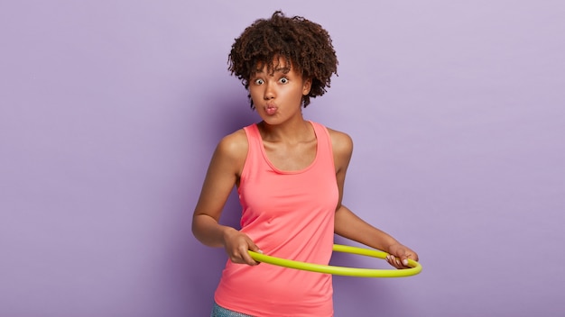 Femme ethnique sportive avec une coiffure frisée garde les lèvres arrondies, fait pivoter le cerceau, est en bonne forme, porte un gilet rose décontracté, a un corps athlétique