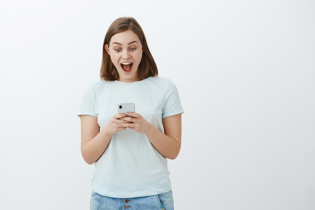 Femme étonnée comme au paradis du message reçu. Fille mignonne enthousiaste en t-shirt souriant se réjouissant, triomphant de bonnes nouvelles en lisant un article intéressant sur un smartphone en regardant l'écran du téléphone portable