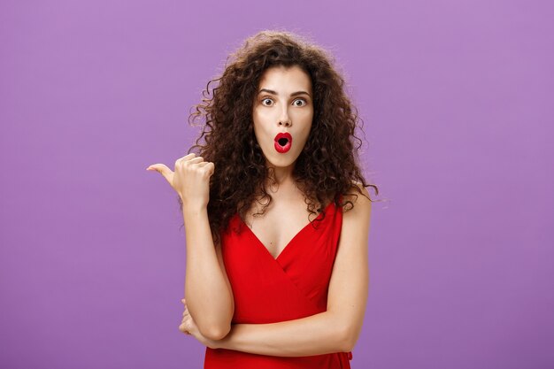 Femme étonnée curieuse et interrogée avec une coiffure frisée en robe rouge élégante pliant les lèvres dans un son wow pointant vers la gauche avec le pouce posant des questions sur une scène époustouflante et passionnante sur un mur violet.
