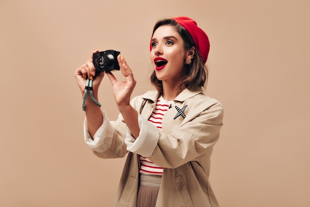 Femme étonnée en tenue tient la caméra sur fond isolé. Belle fille en béret rouge vif fait photo sur fond beige.