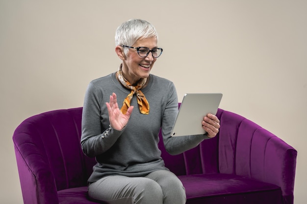 Femme Européenne D'âge Moyen Assise Sur Un Canapé Violet à L'aide D'une Tablette Pour Un Appel Vidéo Et En Agitant