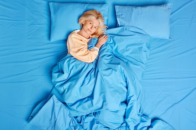 Une femme européenne d'âge moyen calme se réveille satisfaite après avoir vu de bons rêves pose bien dormi sous une couverture bleue porte un pyjama se sent à l'aise profite d'une journée de farniente. Heure du coucher et concept de matin confortable