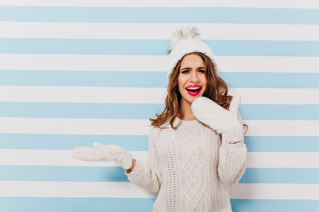 Femme européenne aux cheveux noirs inspirée dans un joli pull blanc posant. Modèle féminin de rêve en vêtements d'hiver en laine, debout sur un mur rayé clair.