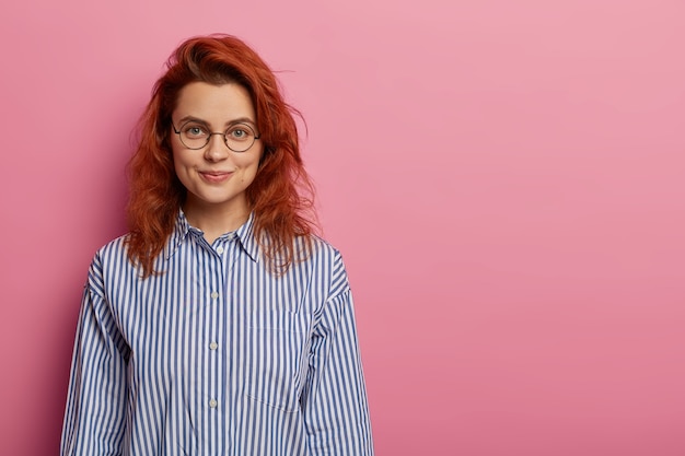 Femme européenne aux cheveux roux optimiste calme regarde directement la caméra, a une expression faciale détendue, un sourire charmant