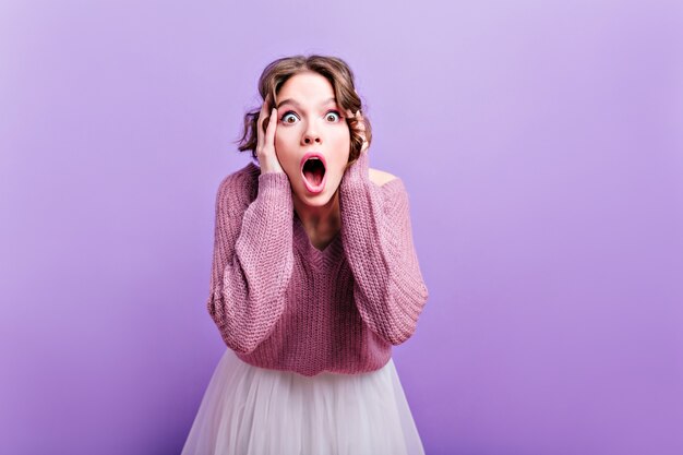 Femme européenne aux yeux sombres posant émotionnellement avec la bouche ouverte. fille aux cheveux bruns surpris avec une coupe courte touchant sa tête pendant une séance photo sur un mur violet.