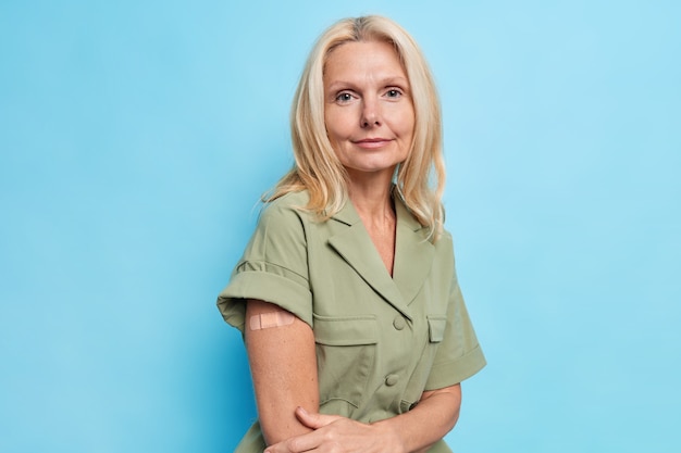 Une femme européenne blonde sérieuse montre un bras vacciné après l'injection de vaccin porte des poses de dess contre le mur bleu