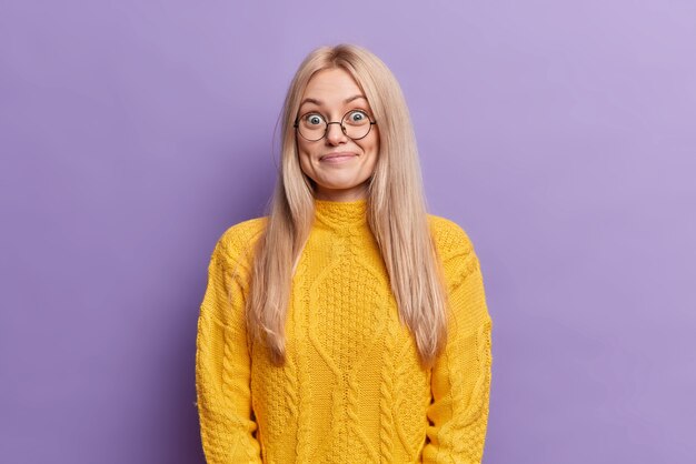 Une femme européenne blonde surprise a une expression joyeuse porte des lunettes rondes entend des nouvelles agréables inattendues vêtues d'un pull jaune