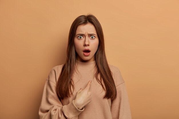 Une femme européenne brune stupéfaite se montre avec une expression stupéfaite indignée, choquée d'être choisie, fronce les sourcils