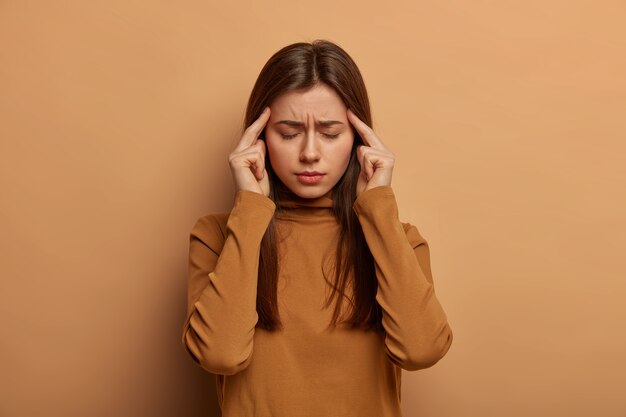 Une femme européenne déprimée et frustrée essaie de se concentrer, garde l'index sur les tempes, réfléchit à une décision importante, ferme les yeux, porte un col roulé marron