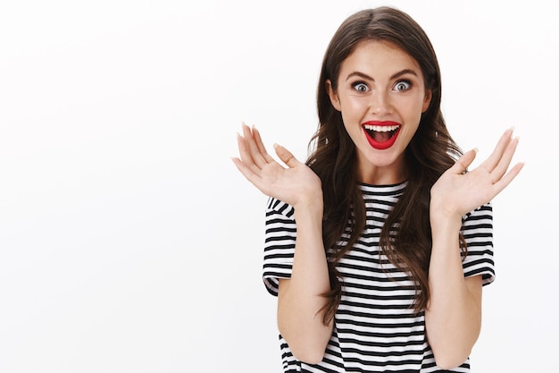 Femme européenne enthousiaste surprise en t-shirt rayé, le rouge à lèvres rouge a l'air étonné et amusé, levant les mains en faisant des gestes ravis d'entendre des nouvelles géniales, de réagir à de merveilleuses nouvelles, mur blanc