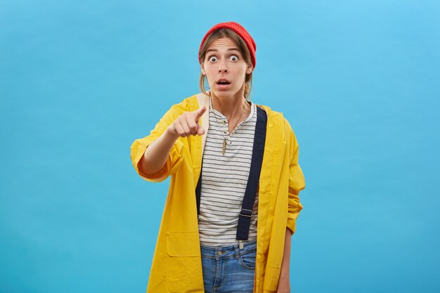 Femme européenne avec une expression surprise vêtue d'une veste jaune décontractée pointant avec l'index en gardant la bouche grande ouverte étant choquée par ce qu'elle voit. Expressions faciales
