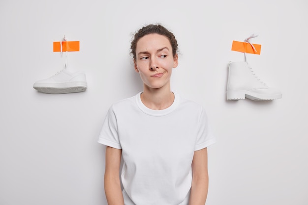 Photo gratuite une femme européenne hésitante porte des lèvres a une expression désemparée sent le doute porte un t-shirt décontracté pose sur fond blanc avec une botte plâtrée et une basket pense quelles chaussures mieux choisir