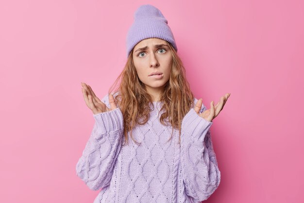 Une femme européenne indignée et perplexe aux cheveux ondulés écarte les mains mord les lèvres ne sait pas quoi faire a une expression désemparée porte un chandail tricoté et un chapeau pose sur fond rose. je m'en fiche