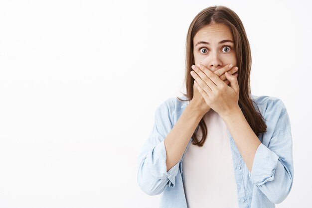 Femme européenne inquiète stupéfaite en chemise bleue sur t-shirt haletant couvrant la bouche avec les paumes des yeux éclatants d'étonnement