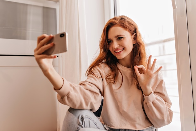Femme européenne inspirée dans des vêtements confortables faisant selfie. Portrait intérieur d'une jolie fille au gingembre.