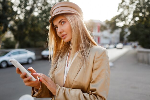 Femme européenne à la mode réussie dans une tenue décontractée élégante posant un téléphone mobile en plein air. Couleurs du coucher du soleil.