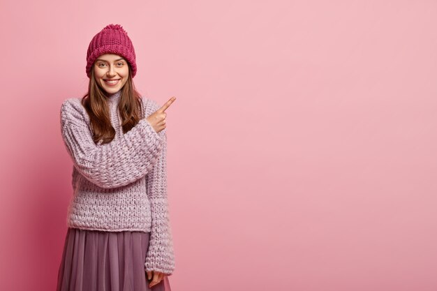 Une femme européenne optimiste pointe dans le coin supérieur droit, porte des vêtements d'hiver à la mode, sourit doucement, fait de la publicité pour quelque chose, isolée sur un mur rose. Regardez l'espace vide pour la promotion