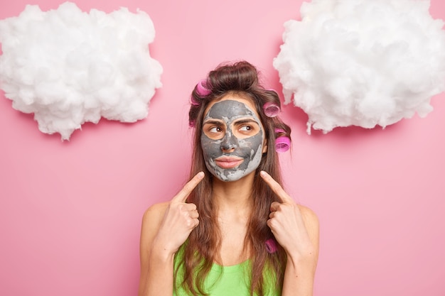 Photo gratuite une femme européenne pointe sur le visage montre que le produit de beauté s'applique sur le visage rend la coupe de cheveux bouclée concentrée de côté isolé sur mur rose