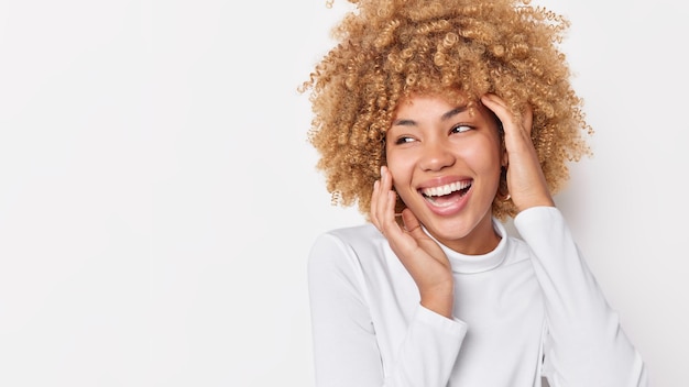 Une femme européenne positive aux cheveux bouclés touche le visage regarde doucement avec bonheur porte un pull décontracté se sent heureux d'avoir une expression heureuse sans soucis pose sur fond blanc avec espace de copie