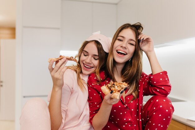 Femme européenne en riant dans des vêtements de nuit mignons, passer la matinée avec sa soeur en masque pour les yeux. Jolies dames blanches mangeant une pizza au fromage avec plaisir.