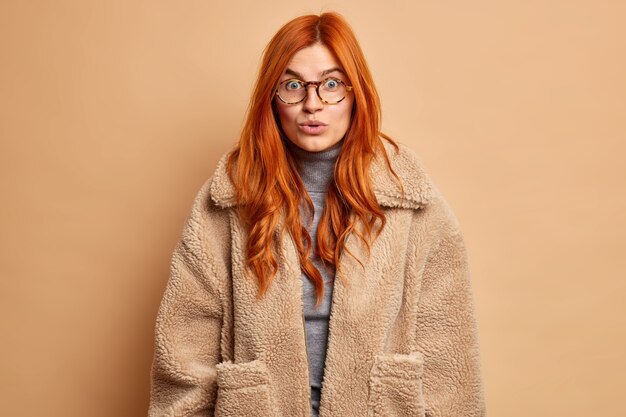 Une femme européenne rousse étonnée entend des rumeurs incroyables ou un secret impressionné par quelque chose porte des lunettes transparentes et un manteau brun fourrure.