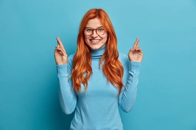 Une femme européenne rousse joyeuse sourit joyeusement croise les doigts fait espoir de recevoir des résultats positifs vêtue d'un col roulé décontracté.