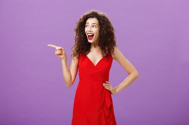 Une femme européenne séduisante amusée avec un maquillage de soirée en robe rouge luxueuse et une coiffure frisée riant à haute voix divertie et heureuse pointant et regardant à gauche intriguée sur fond violet.