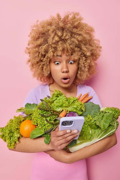 Une femme européenne surprise et choquée aux cheveux bouclés pose avec des produits d'épicerie mange des aliments sains utilise un smartphone vérifie la boîte e-mail vend des légumes sains en ligne se tient à l'intérieur sur fond rose